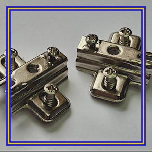 Peut inclure: Gros plan sur deux charnières de meuble métalliques brillantes avec des vis. Les charnières sont rectangulaires avec des bords arrondis et comportent plusieurs trous de vis. Les vis sont argentées et ont une tête en forme de croix. Le fond est gris clair.