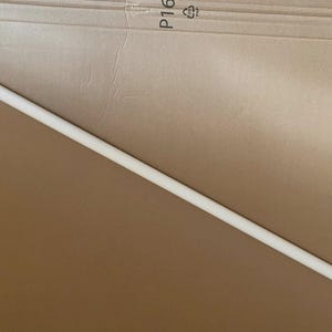 May include: A long, straight, white wooden dowel rod. The dowel is cylindrical and appears to be made of solid wood. It is lying diagonally across a brown cardboard surface.