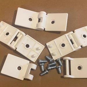 May include: An overhead shot shows several white plastic hinges and a collection of silver screws. The hinges are rectangular with a central hole and a folding mechanism. The screws are small and have a threaded design.