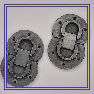 May include: Two gray metal clasps with a circular base and a hinged, rectangular closure. Each clasp has multiple small holes around the perimeter. The clasps are designed to interlock, creating a secure closure mechanism.