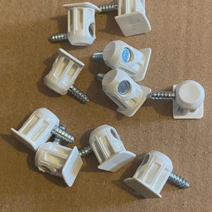 Può includere: Una collezione di connettori per mobili in plastica bianca con viti argentate. I connettori hanno una base quadrata e un corpo cilindrico con un foro per la vite. Le viti sono filettate.