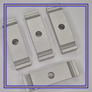 May include: Four rectangular, silver-colored metal pieces with a central hole and grooved edges. The pieces are arranged on a white surface, suggesting a hardware or industrial component.