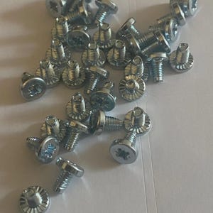 May include: A collection of silver-colored screws with a star-shaped head and a ribbed, circular base. The screws are scattered across a white surface, showcasing their metallic sheen and detailed design. Ideal for hardware and repair projects.