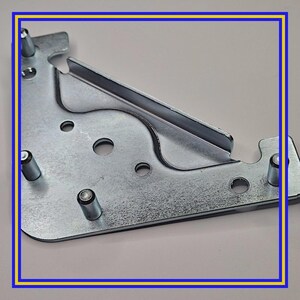 May include: A galvanised metal furniture corner bracket with multiple holes and protruding cylindrical pins. The bracket has a curved design with a slotted edge. The metal has a shiny, reflective surface.