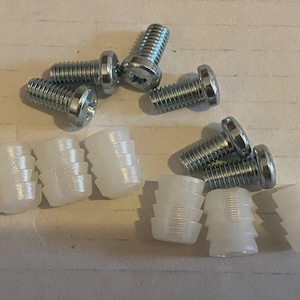May include: A collection of silver metal screws and white plastic dowels. The screws have a flat head and are threaded. The dowels are cylindrical with ridges. The items are arranged on a light-colored surface.