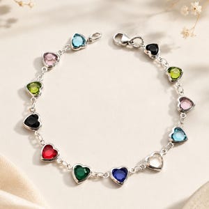 May include: A silver chain bracelet featuring a series of heart-shaped gemstones in various colors, including red, blue, green, black, pink, and light blue, connected by small silver links and secured with a lobster claw clasp.