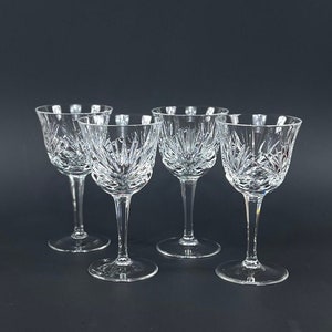 May include: Four clear glass stemmed drinking glasses with a cut crystal design. The glasses are arranged in a row on a black surface.