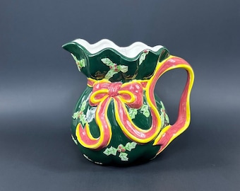 May include: Green ceramic pitcher shaped like a Christmas stocking with a red and yellow bow. The pitcher has a white interior and a handle.