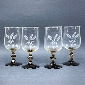 May include: Set of four clear glass drinking glasses with etched wheat designs. The glasses have gold rims and black stems.