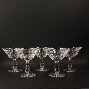 May include: Six clear crystal champagne coupe glasses. Each glass features a wide, shallow bowl and a decorative stem. The glasses are arranged in a row against a dark background, showcasing their intricate cut crystal design.