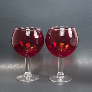 May include: Two clear glass wine glasses filled with a deep red liquid. The glasses have a wide bowl and a long stem.