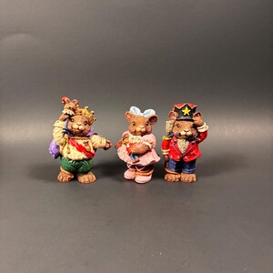 May include: Three decorative mouse figurines. One mouse wears a crown and carries a purple bag. Another mouse wears a pink dress and holds a basket. The third mouse is dressed as a soldier with a red jacket and a tall hat.
