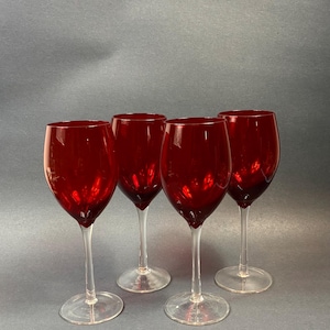 May include: A set of four wine glasses featuring deep red, translucent bowls and clear glass stems and circular bases. The glasses are arranged in a group, showcasing the vibrant crimson color of the glass against a neutral background.