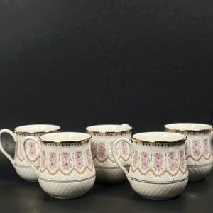 May include: Five white ceramic mugs with gold trim and pink floral patterns. The mugs have a curved handle and a textured base. The mugs are arranged in a group against a dark background.