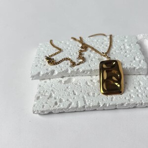 May include: A gold necklace featuring a rectangular pendant with a textured design. The pendant hangs from a fine chain. The necklace is presented on a white, textured surface.