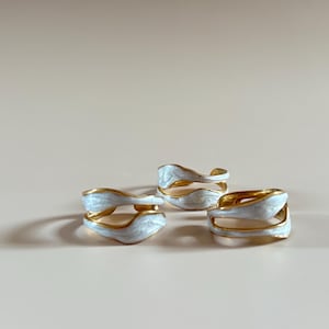 May include: Three white enamel rings with a gold finish. The rings are designed with a wavy, organic shape.