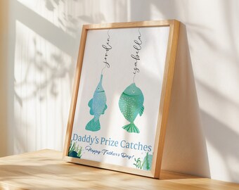 Puede incluir: Una impresión enmarcada con fondo blanco y dos peces azules y verdes colgando de líneas con los nombres "Jordan" e "Isabella" escritos encima. El texto "Daddy's Prize Catches Happy Father's Day!" está impreso debajo de los peces.