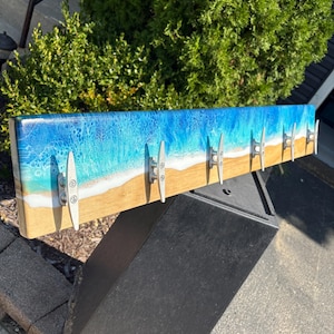 May include: A wooden wall-mounted coat rack with a beach-themed design. The top section features a blue and white resin wave pattern, transitioning to a sandy beach color. Six silver cleat hooks are evenly spaced along the bottom.