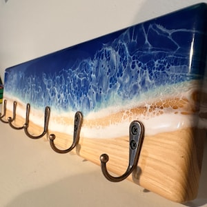 May include: A rectangular wooden wall hook rack featuring a glossy resin art design that mimics an ocean shoreline. The board displays a light wood grain base, a sandy beige strip, and a deep blue sea with white lacy foam waves. Five bronze-toned metal hooks are attached to the front.