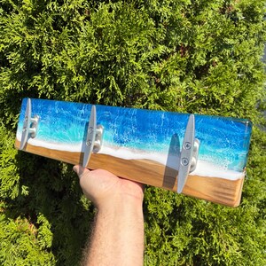 May include: A wooden wall-mounted coat rack with a resin design depicting a beach scene. The resin is blue and white, resembling ocean waves and sand. Three silver cleats are attached for hanging items.