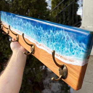 May include: A wooden coat rack with four bronze hooks. The top has a resin design of a blue ocean with white waves and a sandy beach. The wood is light brown. The rack is designed for wall mounting.