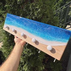 May include: A wooden wall-mounted coat rack with a resin ocean wave design. The rack features a light wood base, white knobs, and silver hooks. The resin art depicts a blue ocean with white foam and a sandy beach.