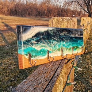 May include: A wooden key holder with five hooks features a resin ocean wave design with shades of blue, green, and white. The key holder is mounted on a wooden fence post.