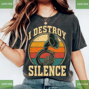 Peut inclure: Un t-shirt gris anthracite avec un graphisme représentant un tuba en laiton centré dans un motif circulaire de coucher de soleil aux rayures jaunes, orange et bleu sarcelle. Le texte "I DESTROY SILENCE" est imprimé en jaune vieilli.
