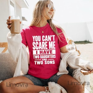 Puede incluir: Una camiseta magenta con el texto blanco "YOU CAN'T SCARE ME I HAVE TWO DAUGHTERS AND TWO SONS". La persona lleva la camiseta al aire libre, sosteniendo un vaso de café helado.