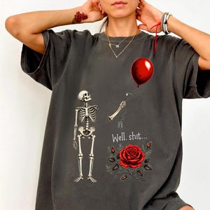 May include: A charcoal grey t-shirt featuring a graphic print of a skeleton, a detached skeletal arm holding a red balloon, a red rose, and the text "Well. shit..." printed in white. The design includes a small spider hanging from the arm.