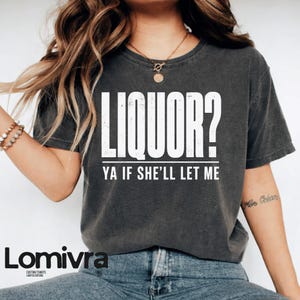 Op de afbeelding: Donkergrijs t-shirt met de witte tekst "LIQUOR? YA IF SHE'LL LET ME". Het shirt heeft een ronde hals en korte mouwen. De merknaam "Lomivra" staat onderaan het shirt.