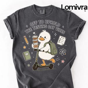 Peut inclure: T-shirt gris foncé avec un canard de dessin animé sur un scooter, tenant du café et un sac à dos. Le texte dit "OFF TO UPHOLD THE TESTING DAY VIBES" avec des illustrations scolaires.