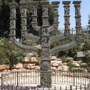 May include: A large bronze menorah sculpture with intricate carvings and Hebrew lettering. The menorah is surrounded by a black metal fence and is set against a backdrop of green trees and rocks.