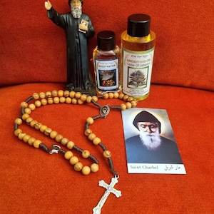 Puede incluir: Un surtido de artículos religiosos sobre una superficie naranja. Incluye una estatua de un santo, dos botellas pequeñas, un rosario con cuentas de madera y una cruz plateada, y una tarjeta con el texto "Saint Charbel".
