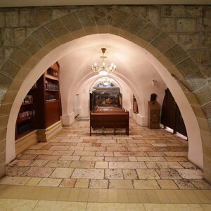 May include: A stone archway leads to a room with wooden pews and a bookshelf. The room is lit by a chandelier.