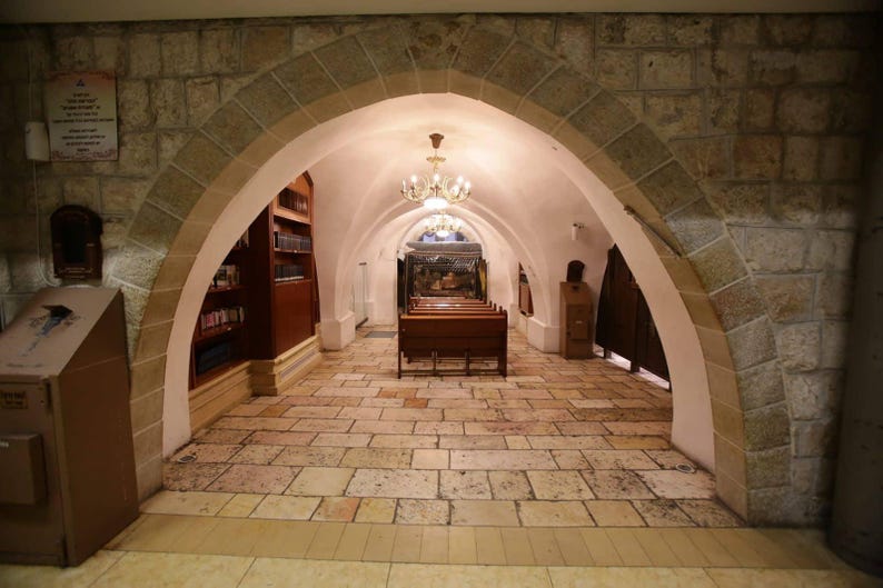 May include: A stone archway leads to a room with wooden pews and a bookshelf. The room is lit by a chandelier.