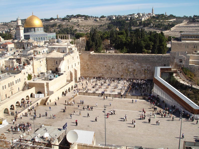 May include: An aerial view of the Western Wall in Jerusalem, Israel. The wall is made of large, rough-hewn stones and is surrounded by a plaza where people are praying. In the background, the Dome of the Rock, a golden-domed mosque, can be seen.
