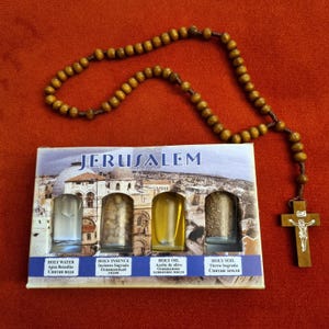 May include: A set of four small glass vials containing holy water, holy incense, holy oil, and holy soil from Jerusalem. The vials are arranged in a row on a white background with the word "JERUSALEM" printed above them. A wooden rosary with a wooden cross is also pictured.