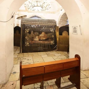 May include: A wooden bench sits in front of a covered shrine with a depiction of a building with a dome. The shrine is in a room with white walls and a tiled floor. A sign on the wall reads "Towards Jerusalem".