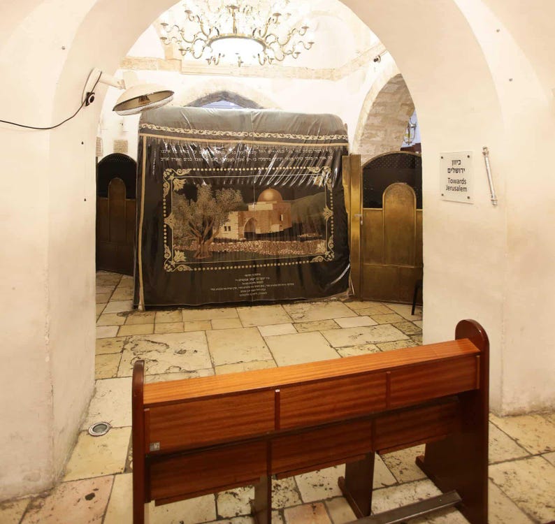 May include: A wooden bench sits in front of a covered shrine with a depiction of a building with a dome. The shrine is in a room with white walls and a tiled floor. A sign on the wall reads "Towards Jerusalem".