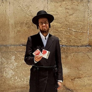 May include: A man wearing a black hat and long black coat stands in front of a stone wall. He is holding two small cards with red hearts on them. The text "Nekudat Chen Haifa 972-54-6818597" is visible at the bottom of the image.