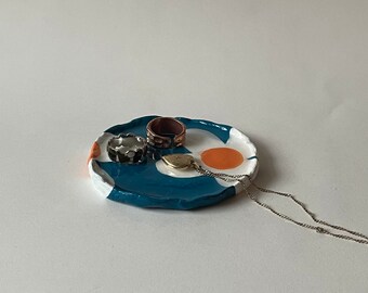 May include: A teal blue ceramic trinket dish with a white and orange design. The dish has a wavy edge and is decorated with a white flower and an orange circle. There are three rings and a necklace on the dish.