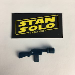 Peut inclure: Un blaster jouet en plastique bleu foncé avec un canon noir. Le blaster est une réplique d'une arme de la franchise Star Wars. L'image comprend également une carte noire avec le texte "STAN SOLO CREATIONS" en jaune.