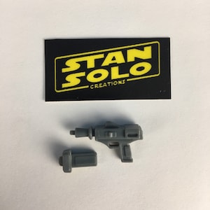 May include: A grey plastic toy blaster with a black handle and a grey magazine. The blaster is from the Star Wars franchise. The image also includes a black card with the text "STAN SOLO CREATIONS" in yellow letters.