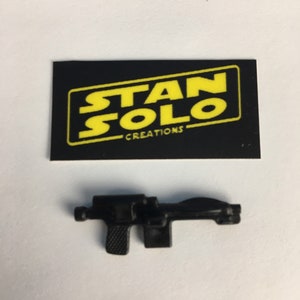 May include: Black plastic toy blaster with a textured grip and a black and yellow "STAN SOLO CREATIONS" logo on a black background.