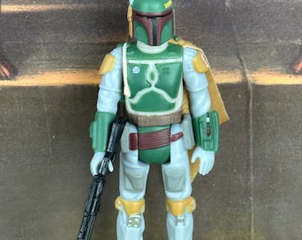 Puede incluir: Figura de acción de Boba Fett, con casco y armadura verdes, traje gris y blanco, y un rifle negro. La figura tiene una capa color canela y está de pie sobre una base amarilla y negra con el texto "STAN SOLO CREATIONS".
