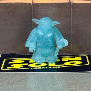 May include: A translucent blue Yoda figurine with a staff. The figure is standing on a black and yellow label that reads "SOLO CREATIONS". The background is a blurred brown and black.