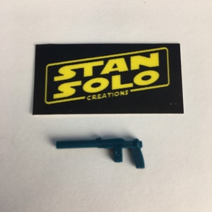 May include: A teal blue plastic toy blaster with a black handle. The blaster is a replica of a weapon from the Star Wars franchise. The image also includes a black card with the text "STAN SOLO CREATIONS" in yellow.