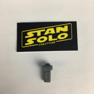 May include: A gray plastic piece with a rectangular shape and a small rectangular opening on the side. The piece is labeled "STAN SOLO CREATIONS".