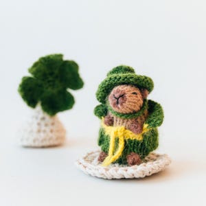 The knitted capybara is shown with the shamrock placed behind it for atmosphere. The soft focus in the background adds depth while keeping the toy as the main subject. The scene feels festive and cozy.